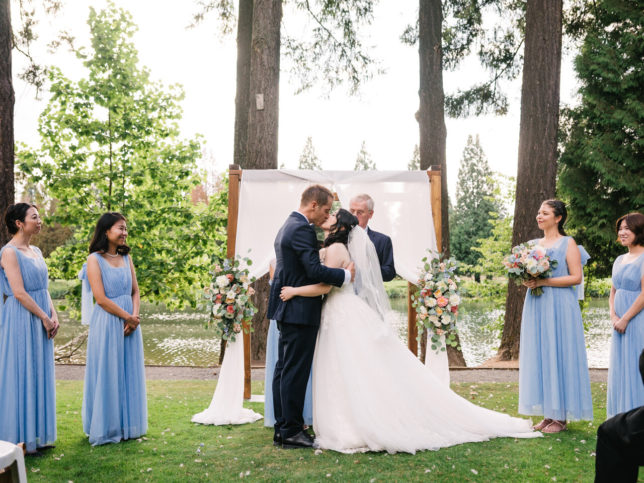  Bride and groom kiss in front of wedding arch before bridesmaids with blue dresses and tall fir trees 