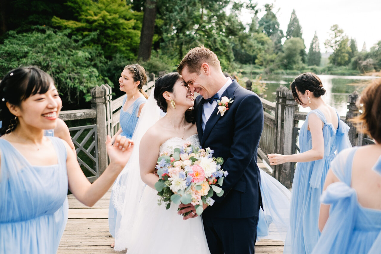  Bride and groom snuggle on wooden bridge while bridesmaids in blue dresses walk around them 