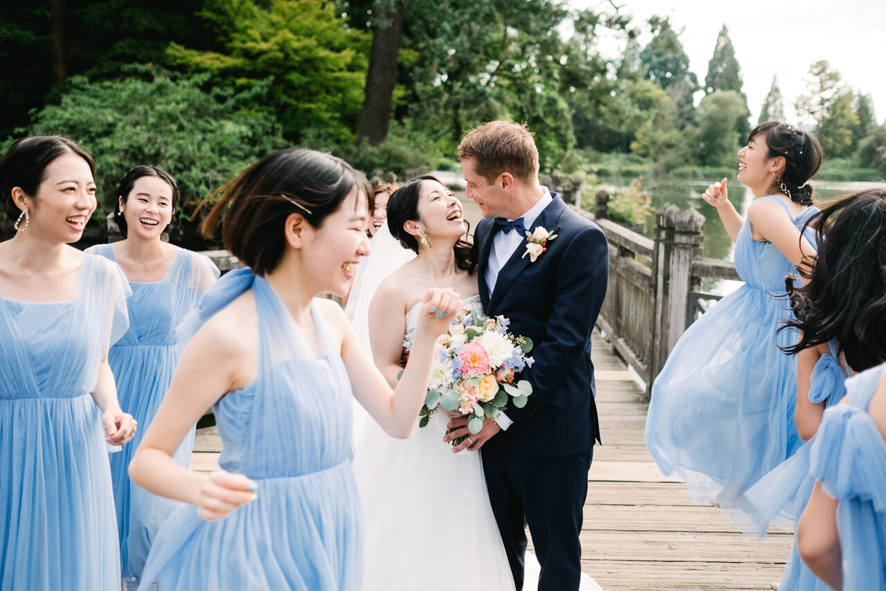  Bride and groom snuggle on wooden bridge while bridesmaids in blue dresses jump around them 