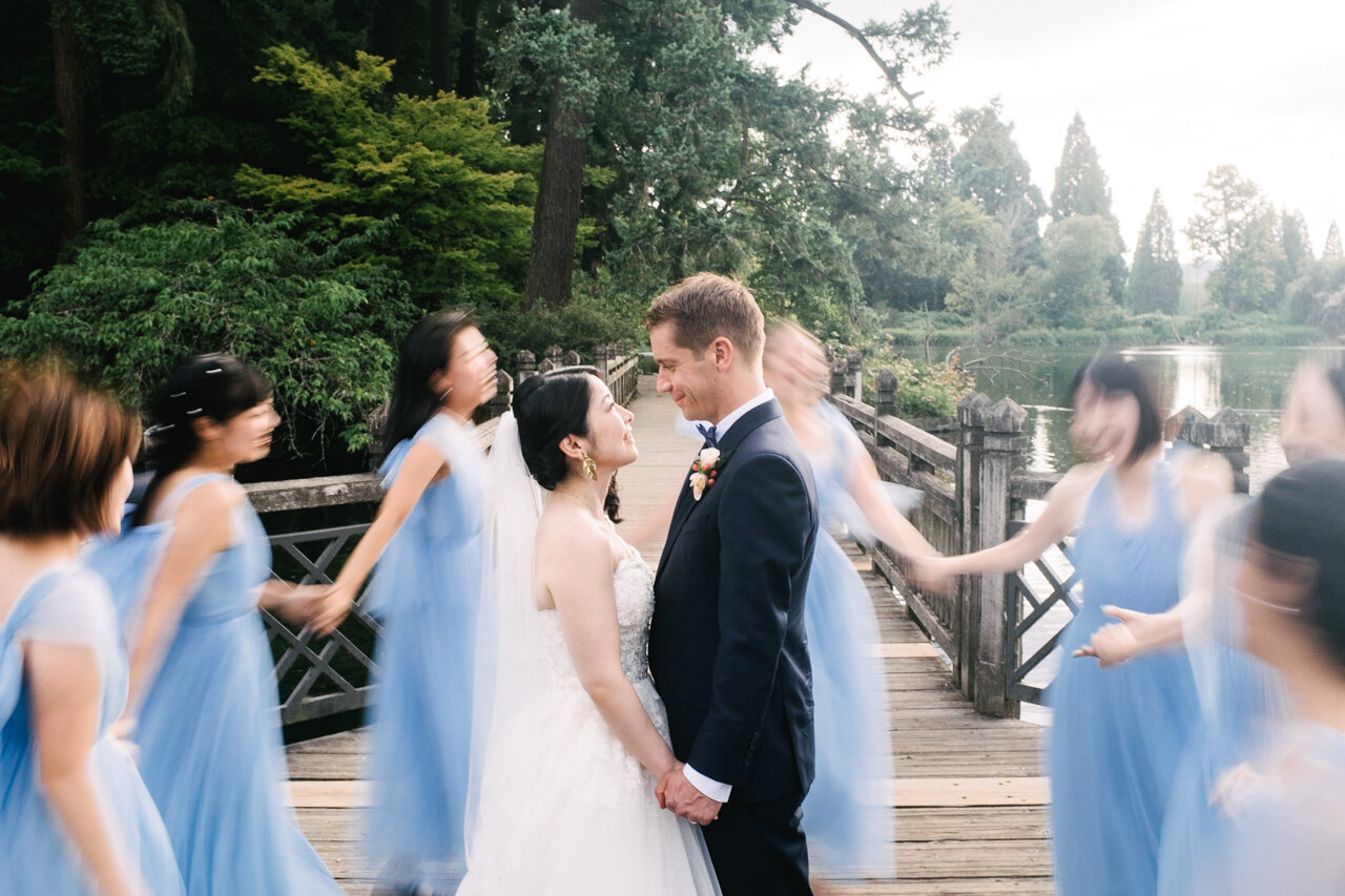  Bride and groom remain still while bridesmaids in blue dresses move in blurry motion around them on wooden bridge walkway 