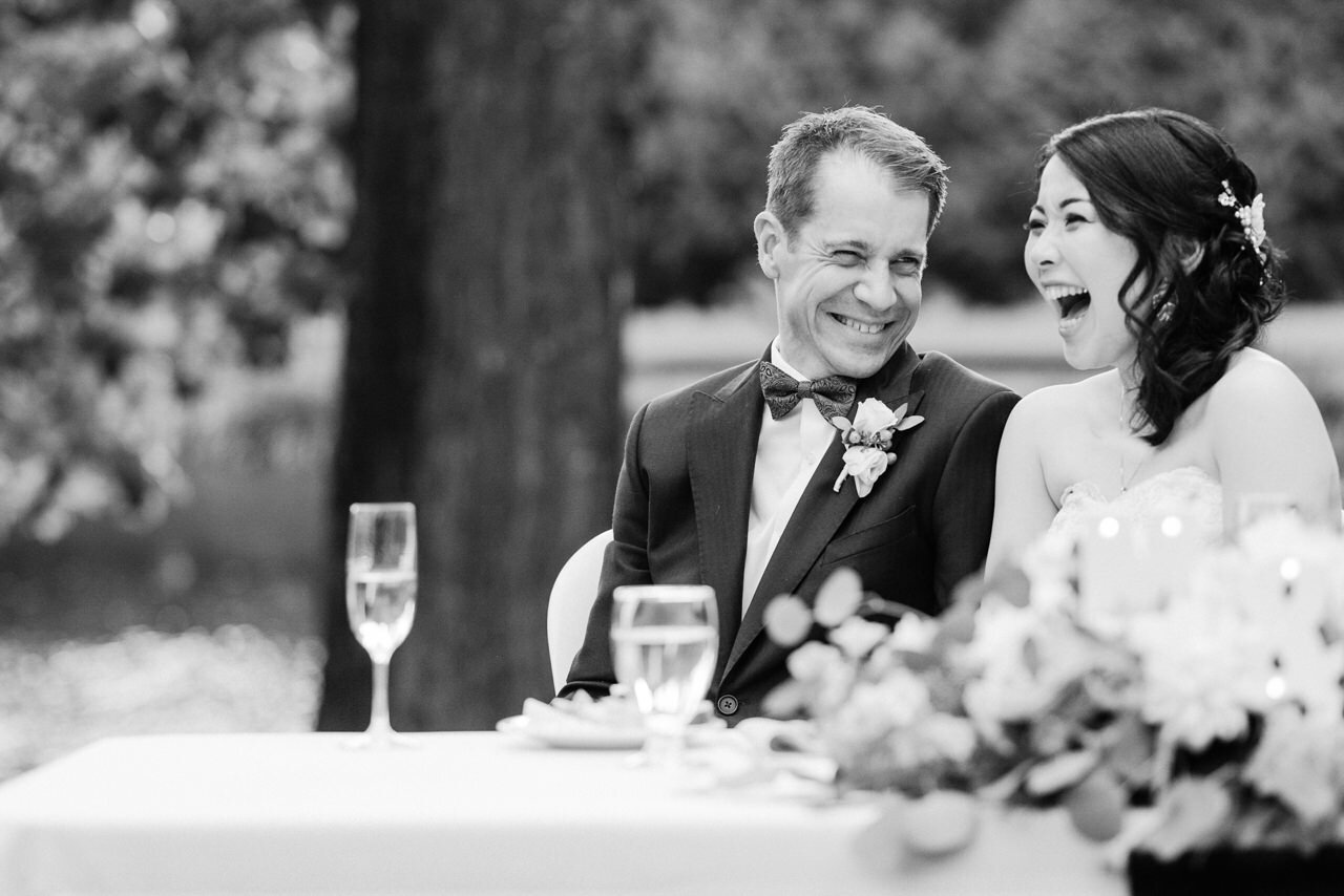  Black and white photo of bride bursting out laughing while groom laughs with her at table 