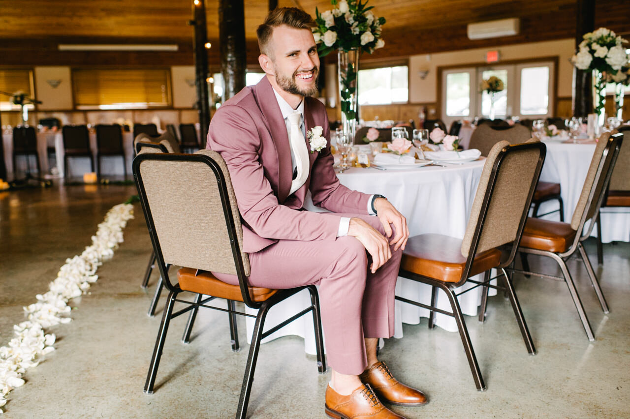  Groom in purple suit sits at reception table laughing with ceremony flowers behind him 