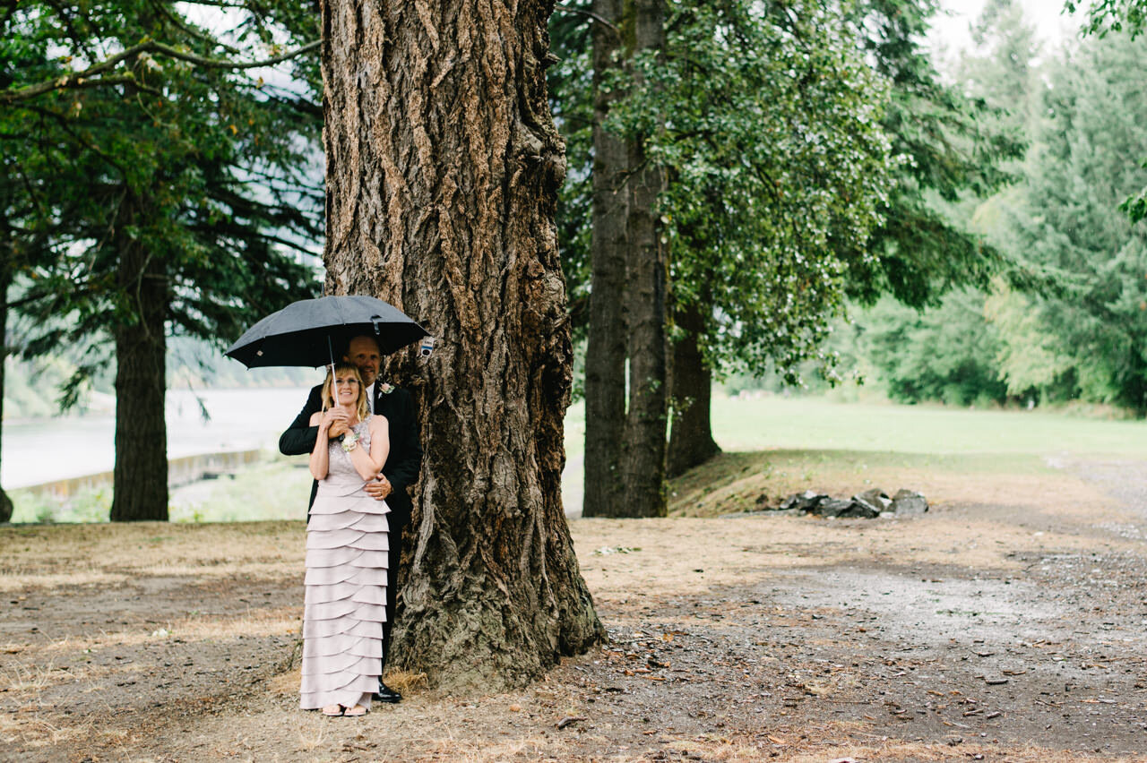 Parents of the groom stand under umbrella by large fir tree while waiting for photos 