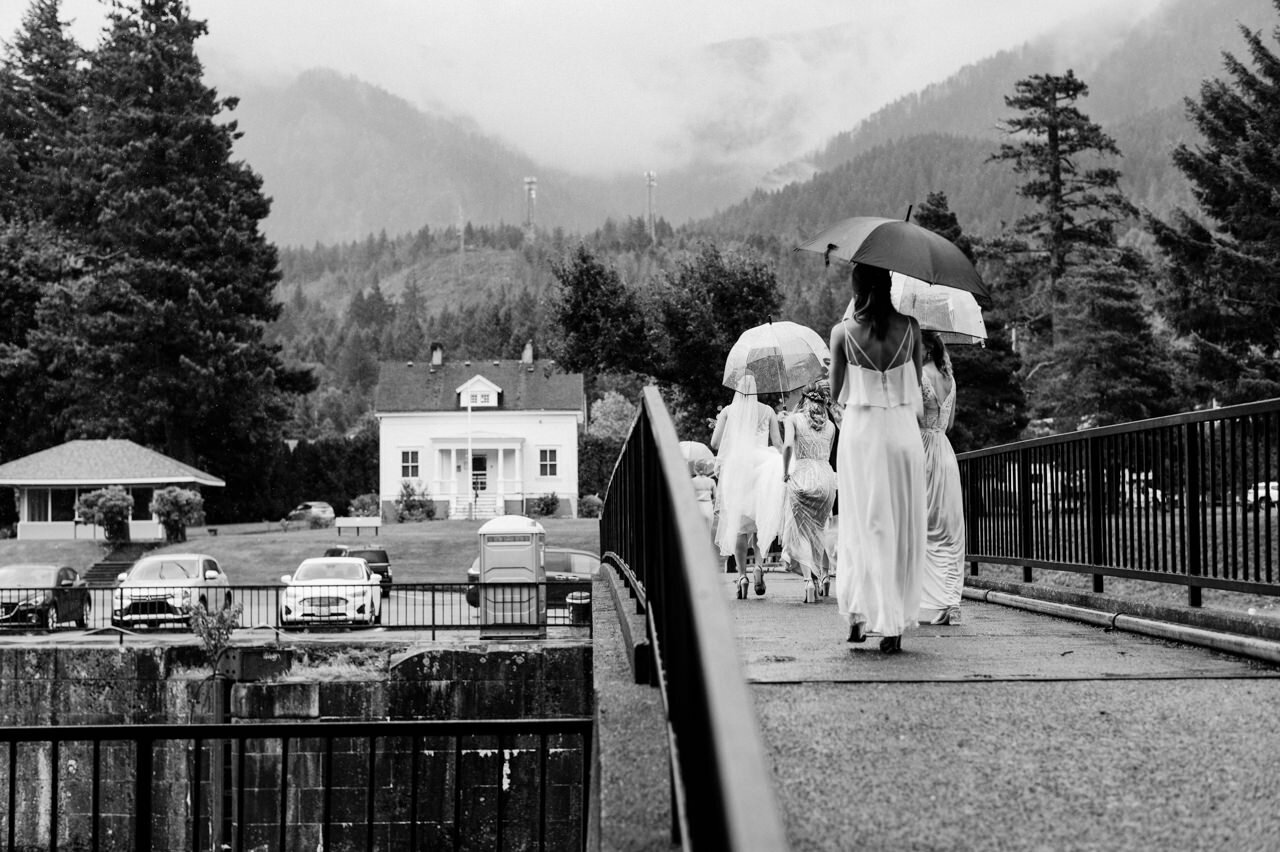  Bride and bridesmaids walk away across cascade locks bridge during rain 