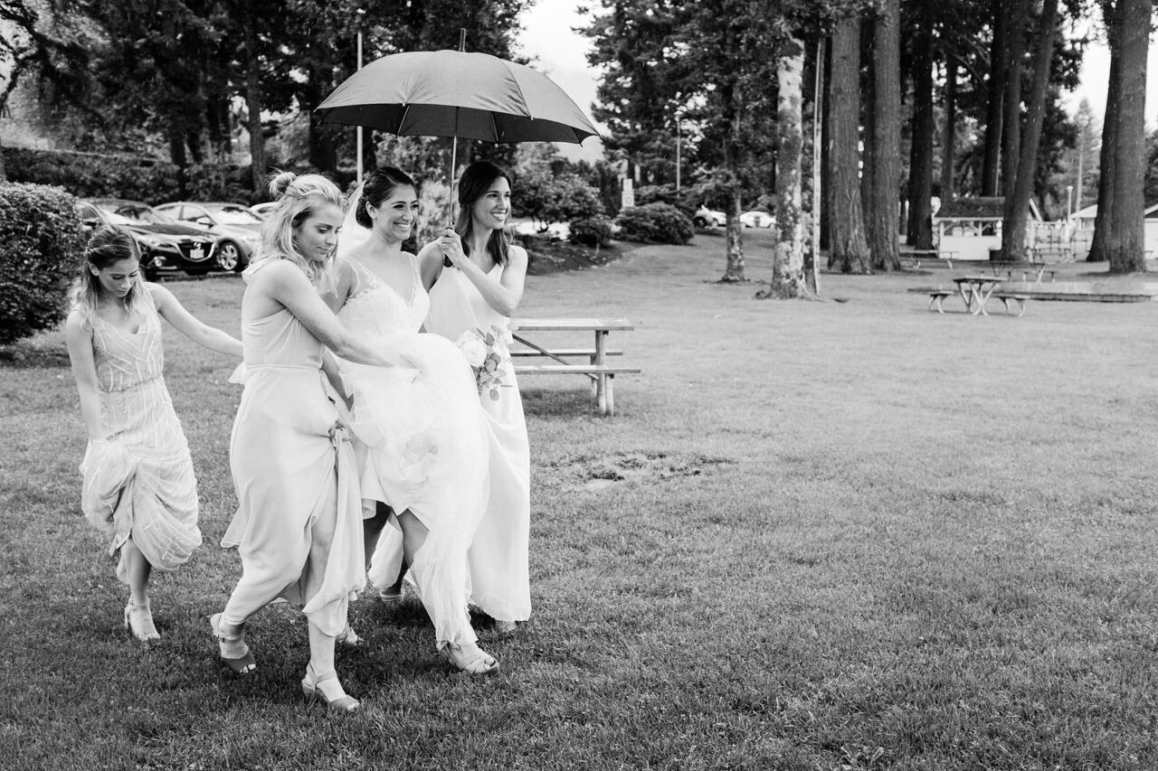  Bride approaches wedding ceremony under umbrellas at cascade locks while bridesmaids help her with dress 