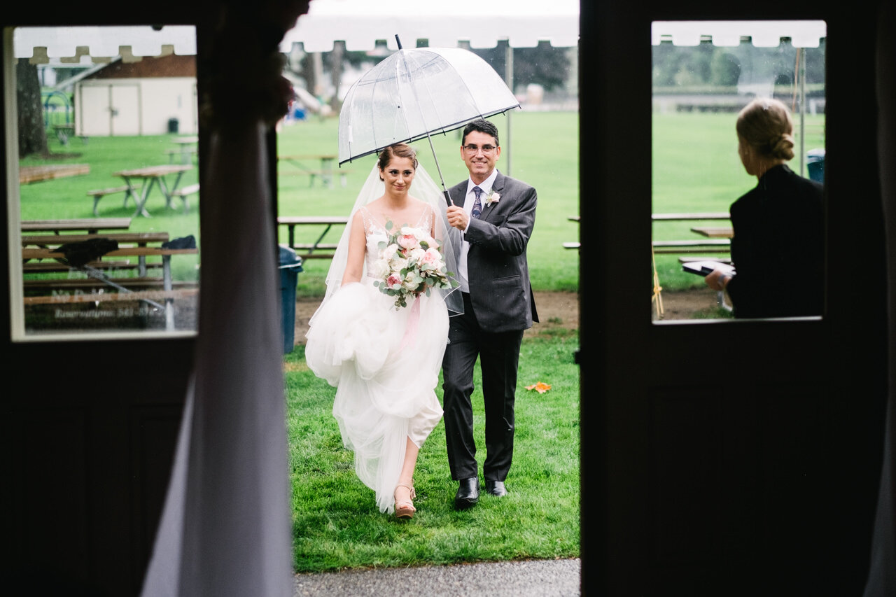  Bride walks with father in rain approaching indoor wedding ceremony 