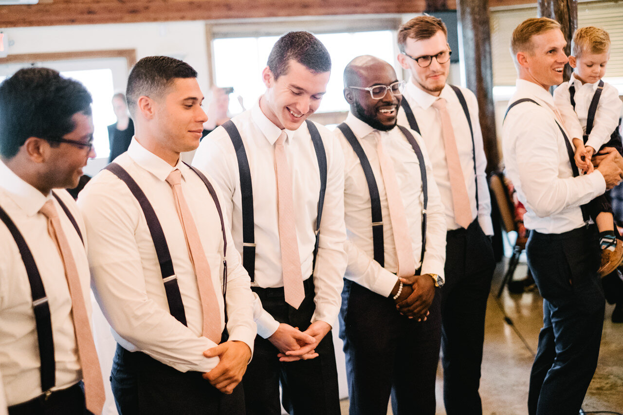  Groomsmen in black suspenders and pink ties laugh in line during wedding 