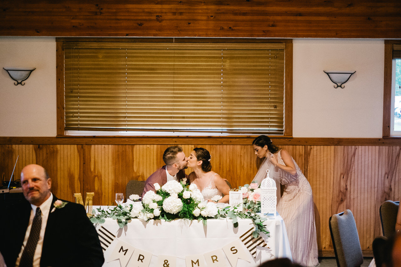  Seated at mr and mrs wedding table, bride and groom kiss while bridesmaid fixes dress 