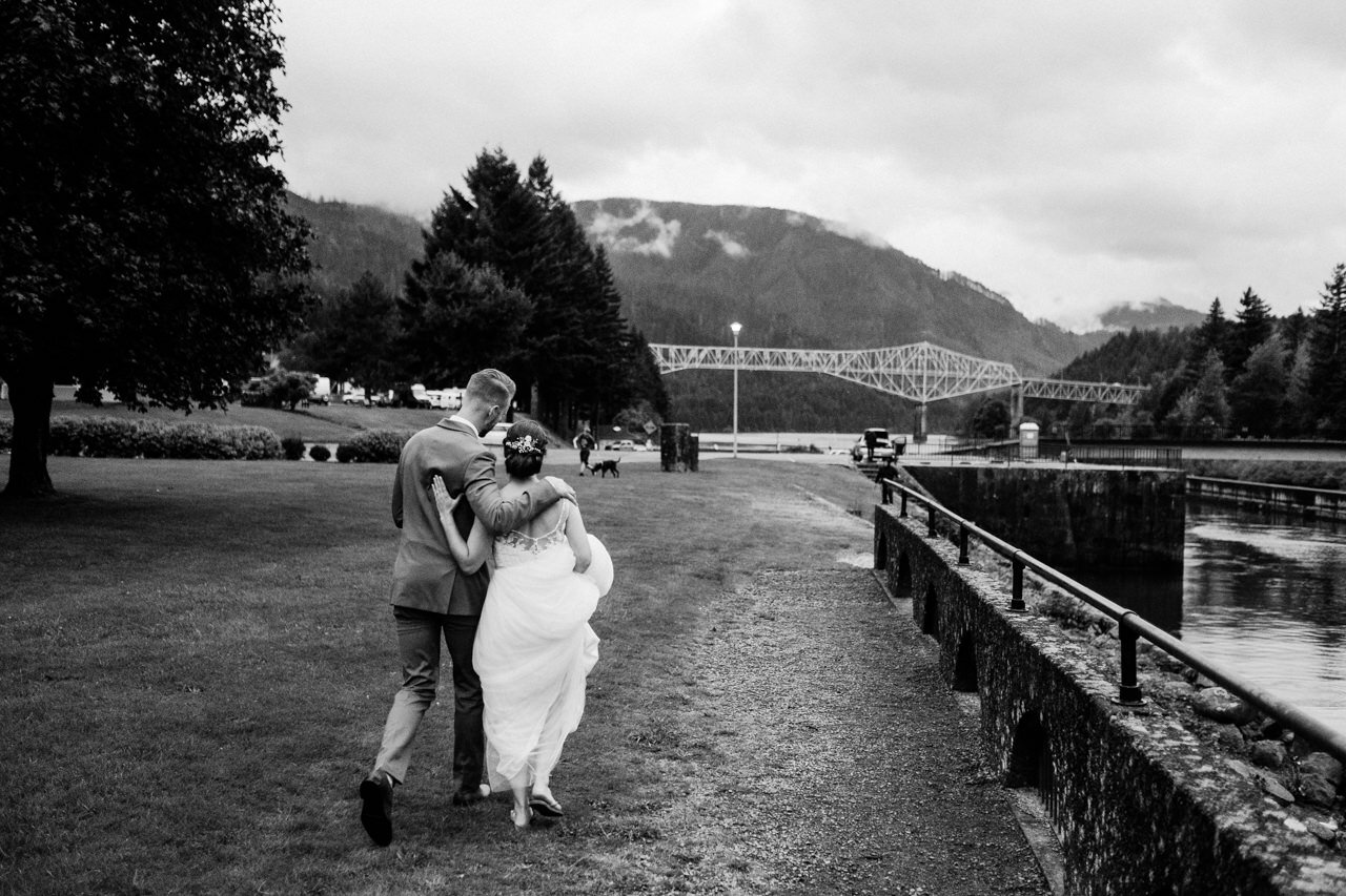  Bride and groom walk with arms around each other towards the bridge of the gods 