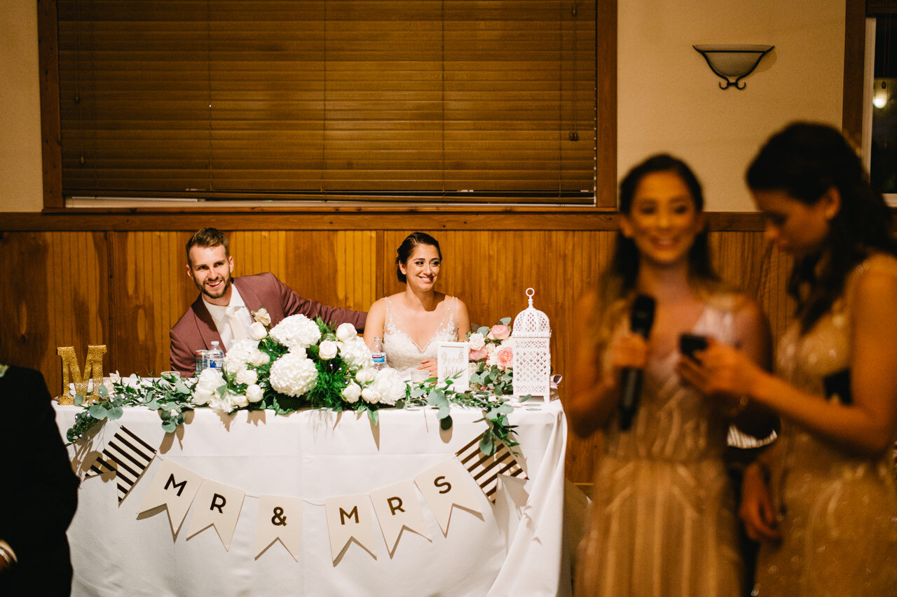  Bride and groom smile together in spotlight during toast by sisters 