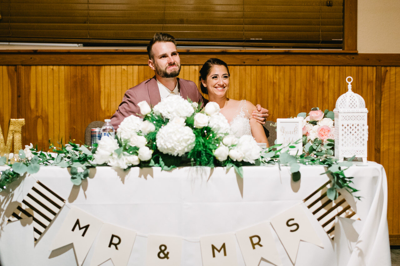  Groom wraps arm around bride seated at wedding table with white hydrangeas and banner mr and mrs 