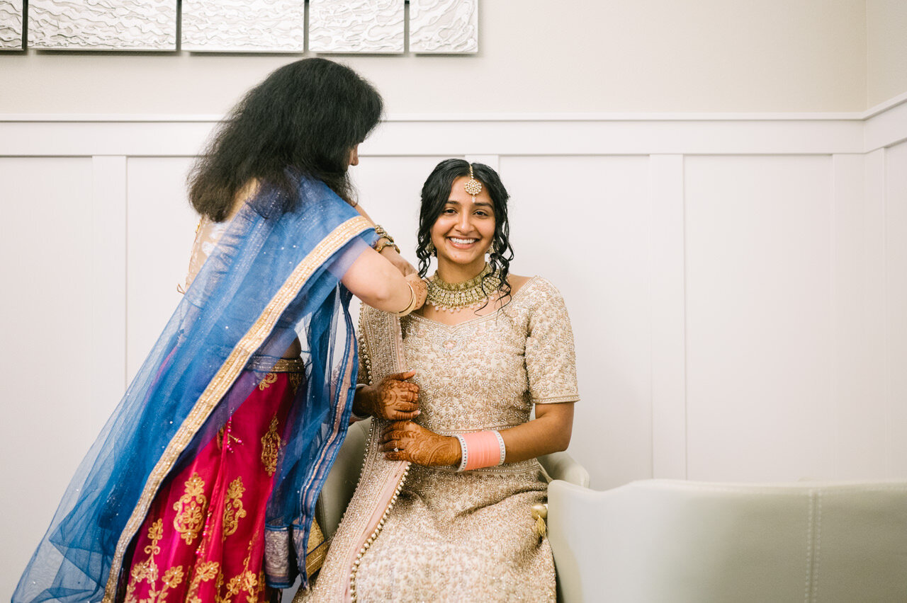  Bride smiles while Indian auntie in blue and red sari adjusts her sari 