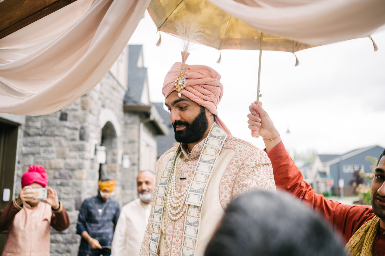  Groom smiles with pink turban and money necklace under umbrella 