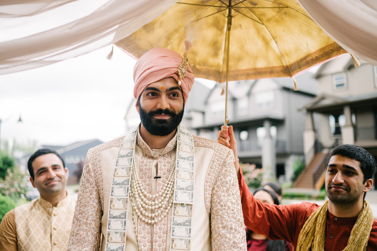  Indian groom smiles with pink turban and cream necklace under umbrella 