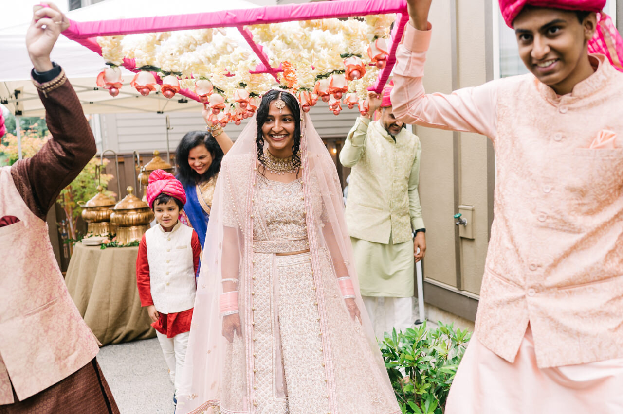  Indian bride walks toward ceremony under pink and cream covering 