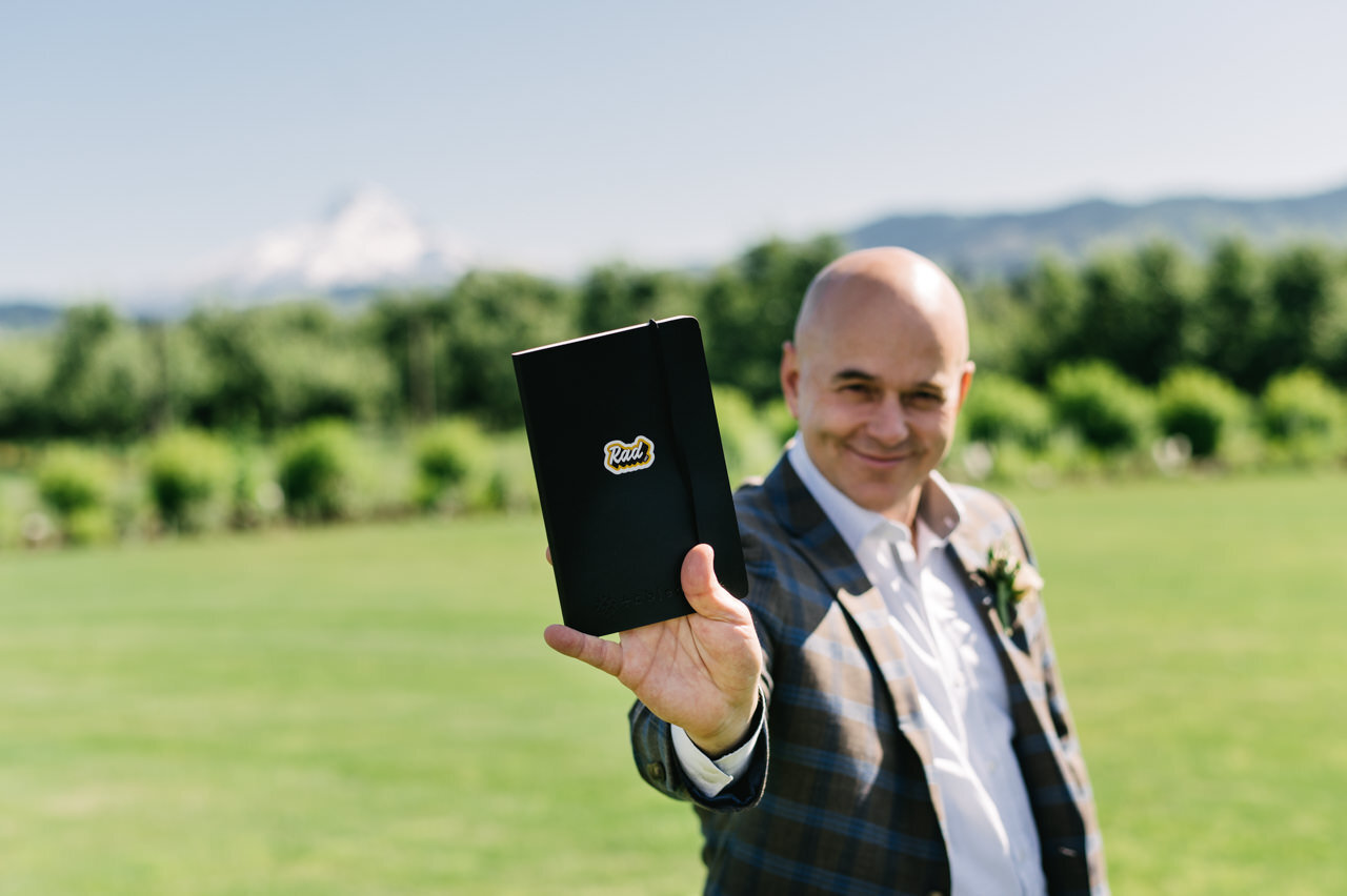  Best man holds notebook for wedding in front of lawn with mt hood 