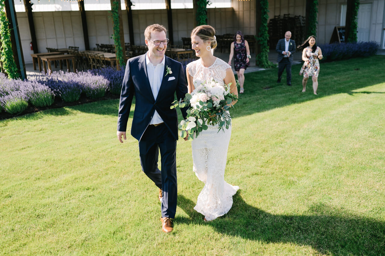  Bride and groom walk on lawn in front of lavender with guests following behind 