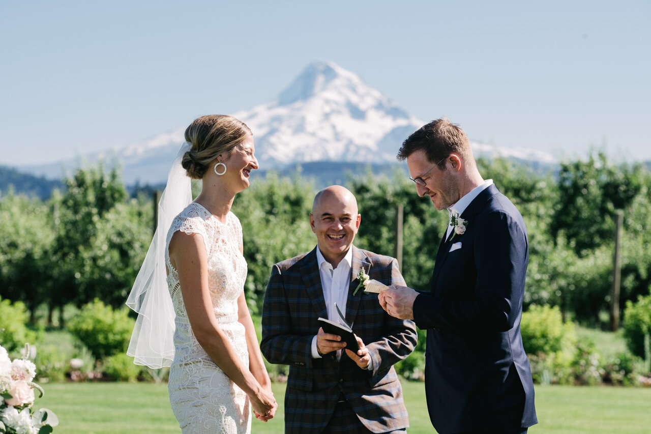  Groom reads bride vows while officiant laughs with mt hood behind them 