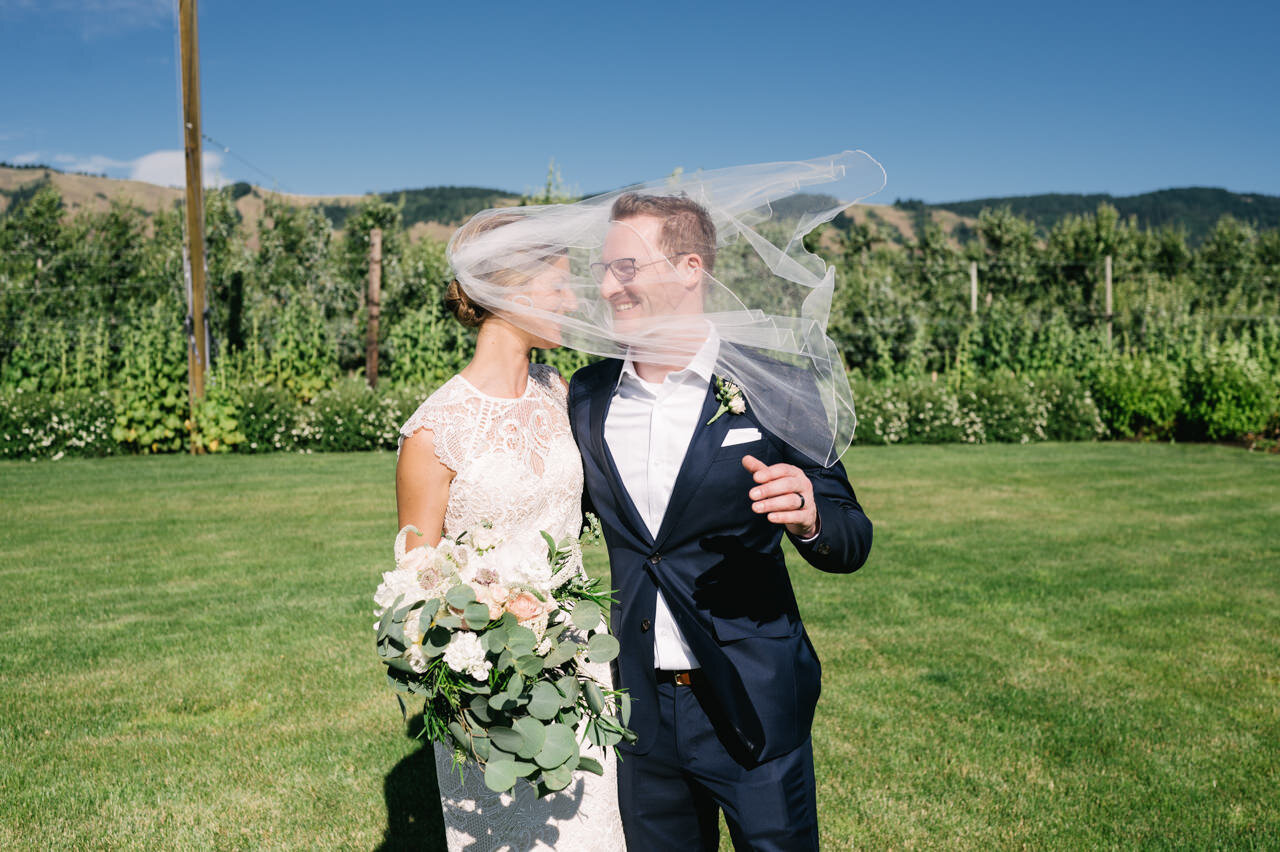  Bride's veil blows in wind across grooms face in front of sunny orchard 