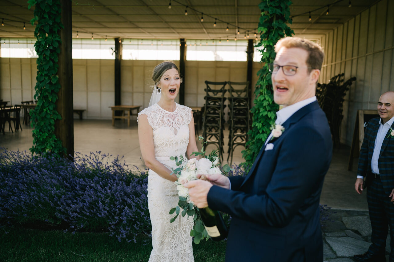  Bride and groom laugh as groom tries to pop champagne in front of pavilion 