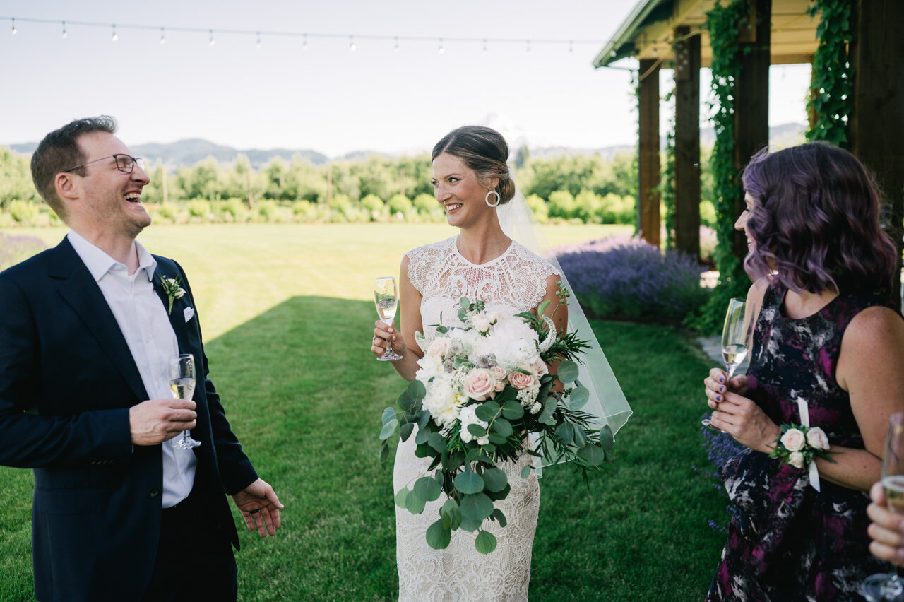  Groom laughs before toast at sunny orchard while bride holds large peach and white bouquet 