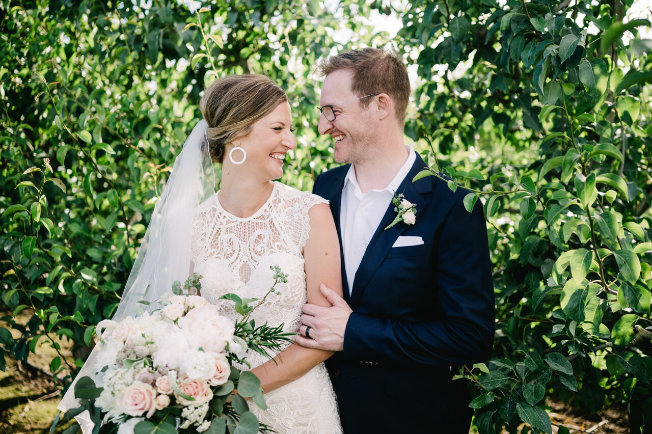  Bride with ornate lace dress and groom smile together in orchard 