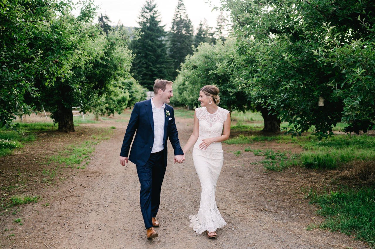  Bride and groom happily walk down dirt road in orchard 