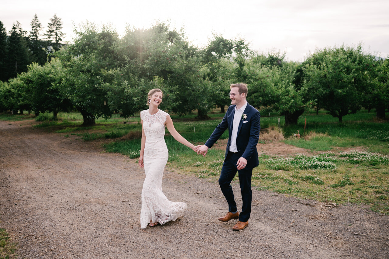  Wedding couple walks across dirt road with orchard behind them 