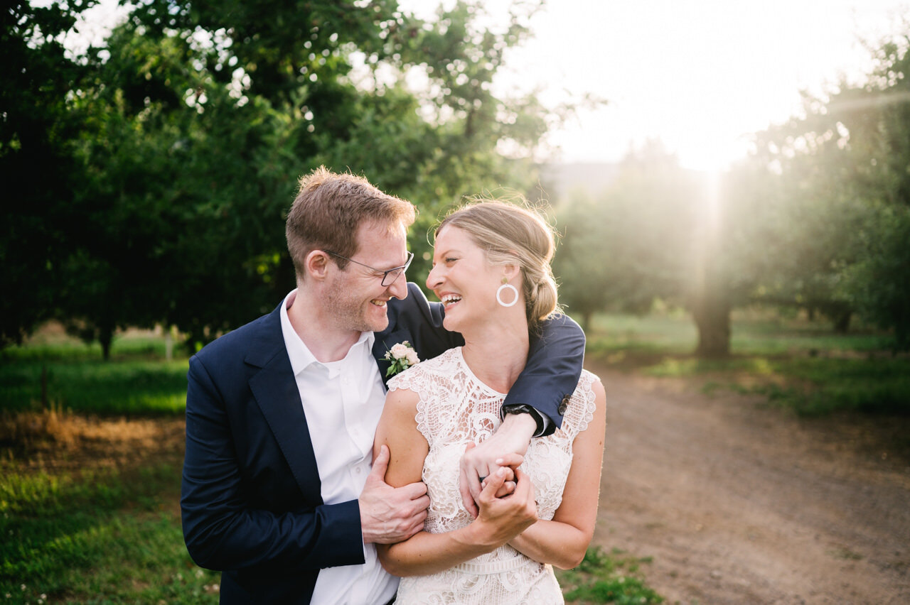  Bride with lace dress and groom laugh in sunlight orchards holding hands 