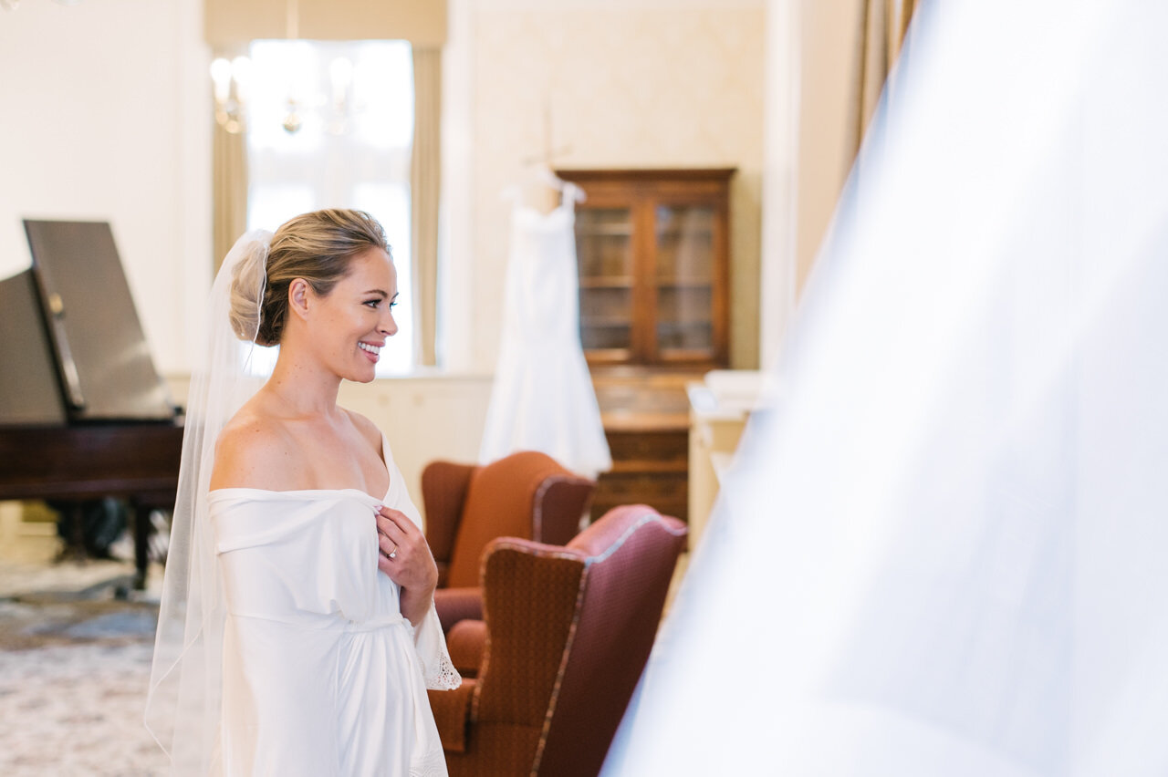  Bride with veil on looking in mirror with dress hanging in the background 