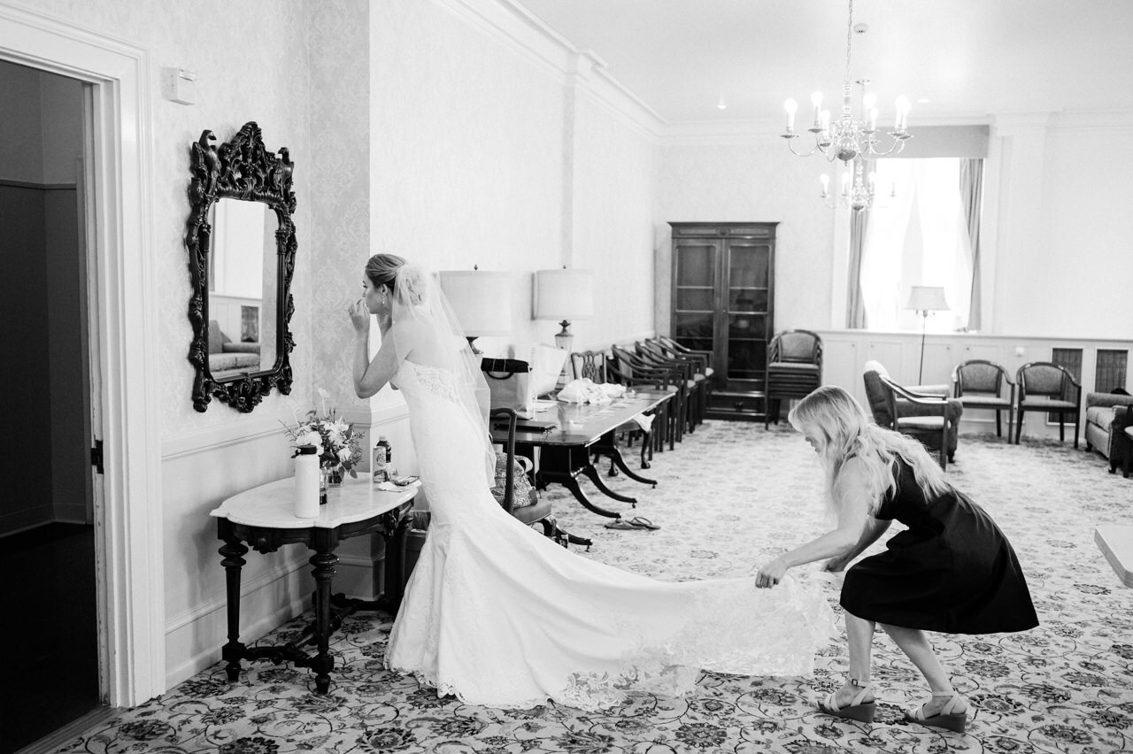  Maid of honor adjusts wedding dress train while bride applies last minute makeup in mirror 