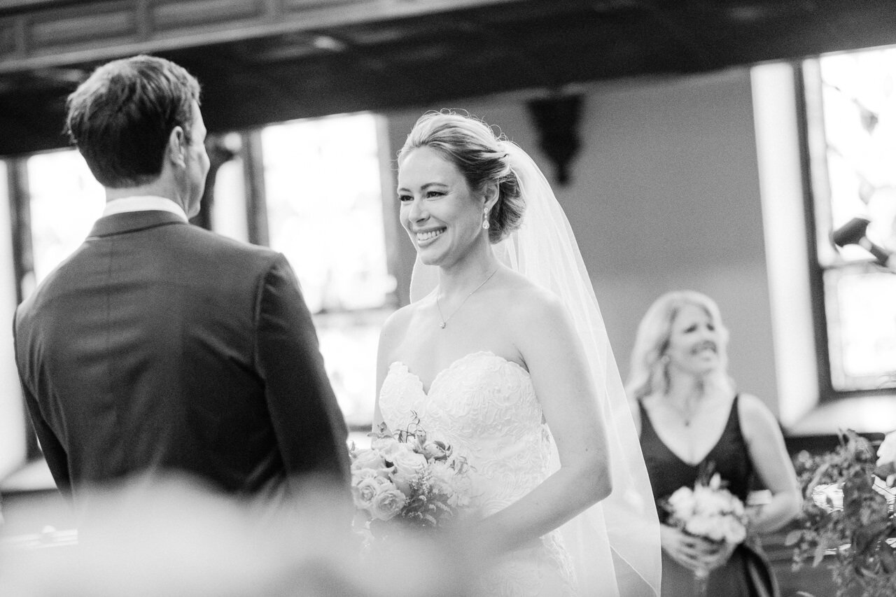  Black and white photograph of bride smiling at groom during ceremony 