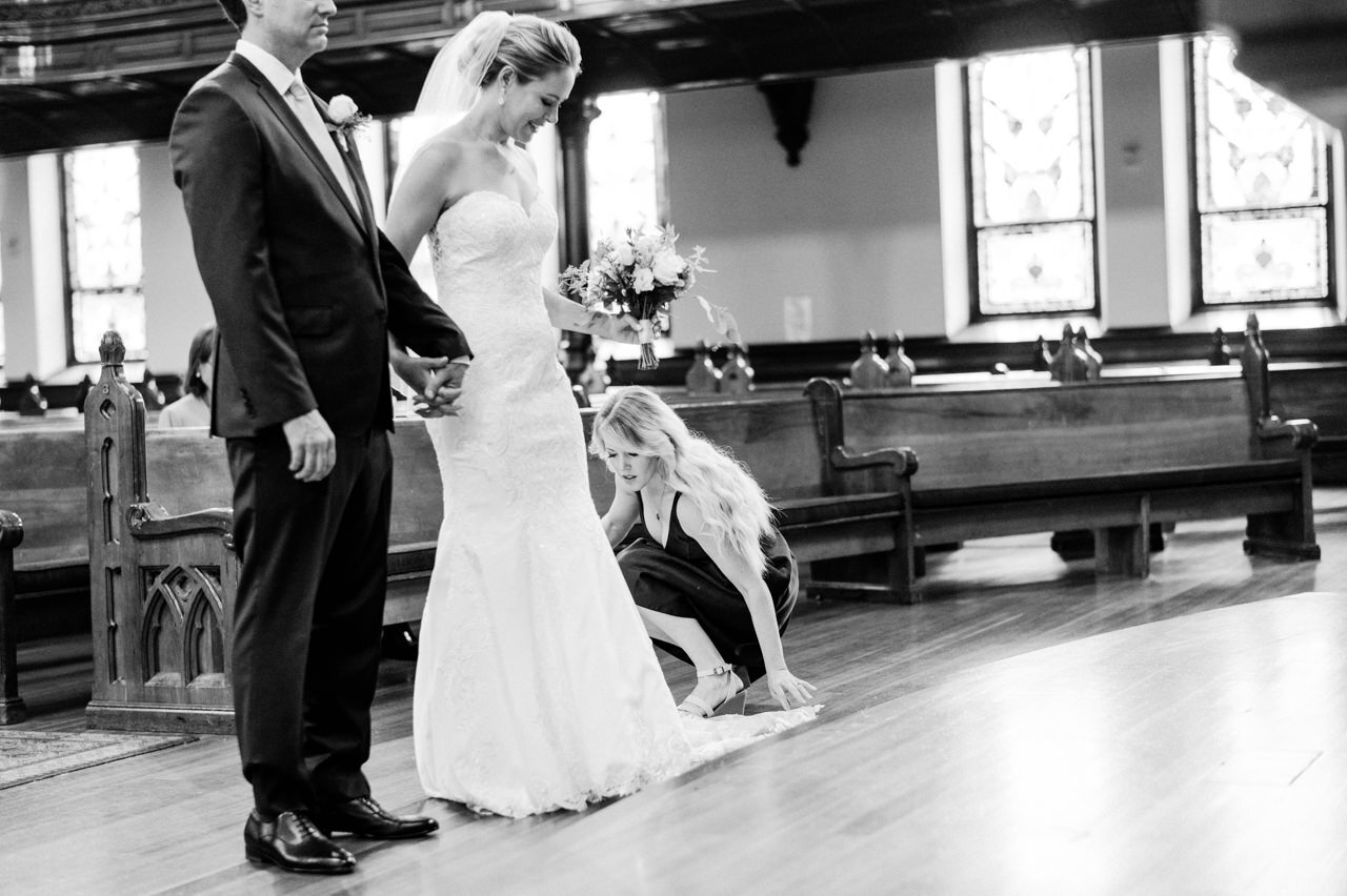  During ceremony, maid of honor adjusts brides train as the bride and groom face the altar 