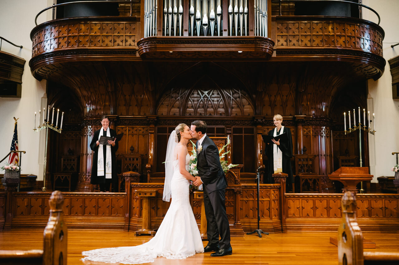 Bride and groom kiss in first presbyterian church with clergy looking on with a pipe organ behind them 
