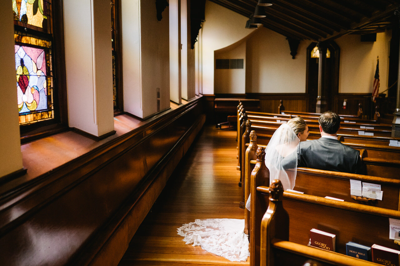  Bride and groom sit in pews of church while light shines through stained glass 