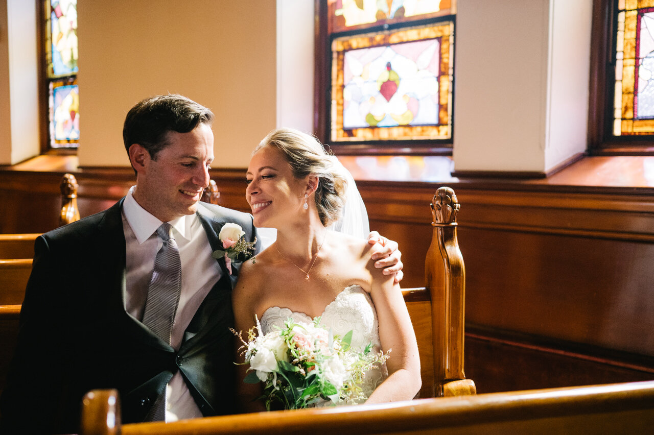  Bride and groom smile at each other in church pews with stained glass behind them 