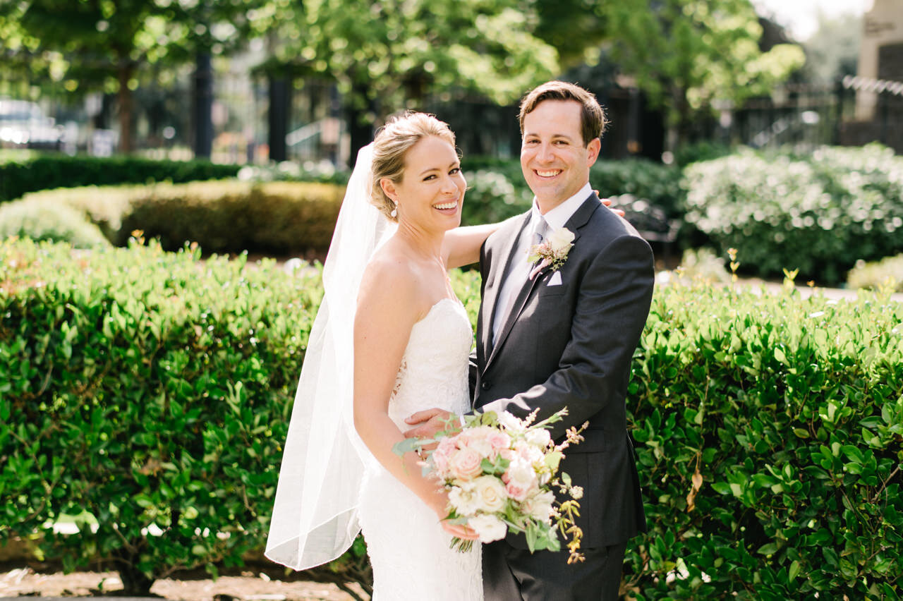  Bride and groom laugh for wedding portrait as she holds light pink and white florals 