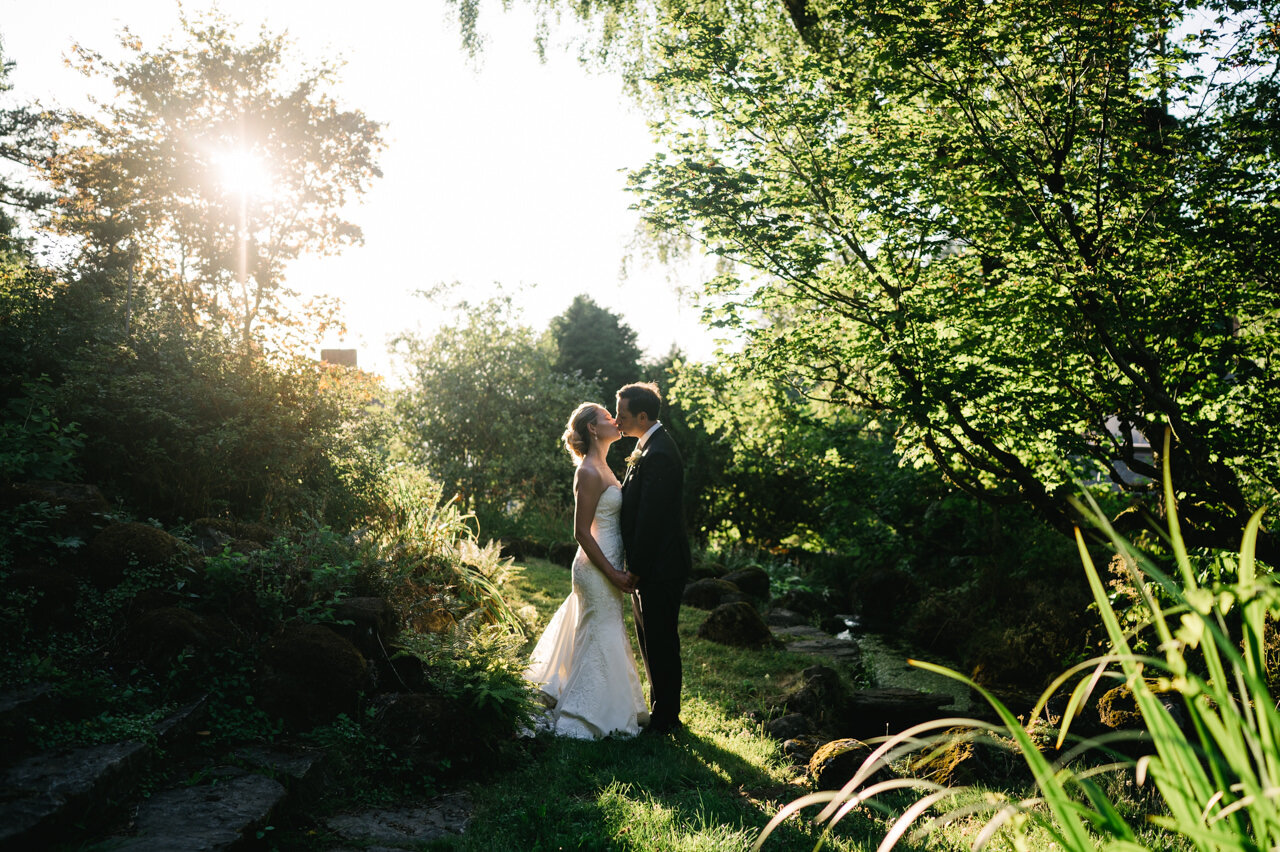  Bride and groom kiss by pond with sunlight behind them in serene grove 