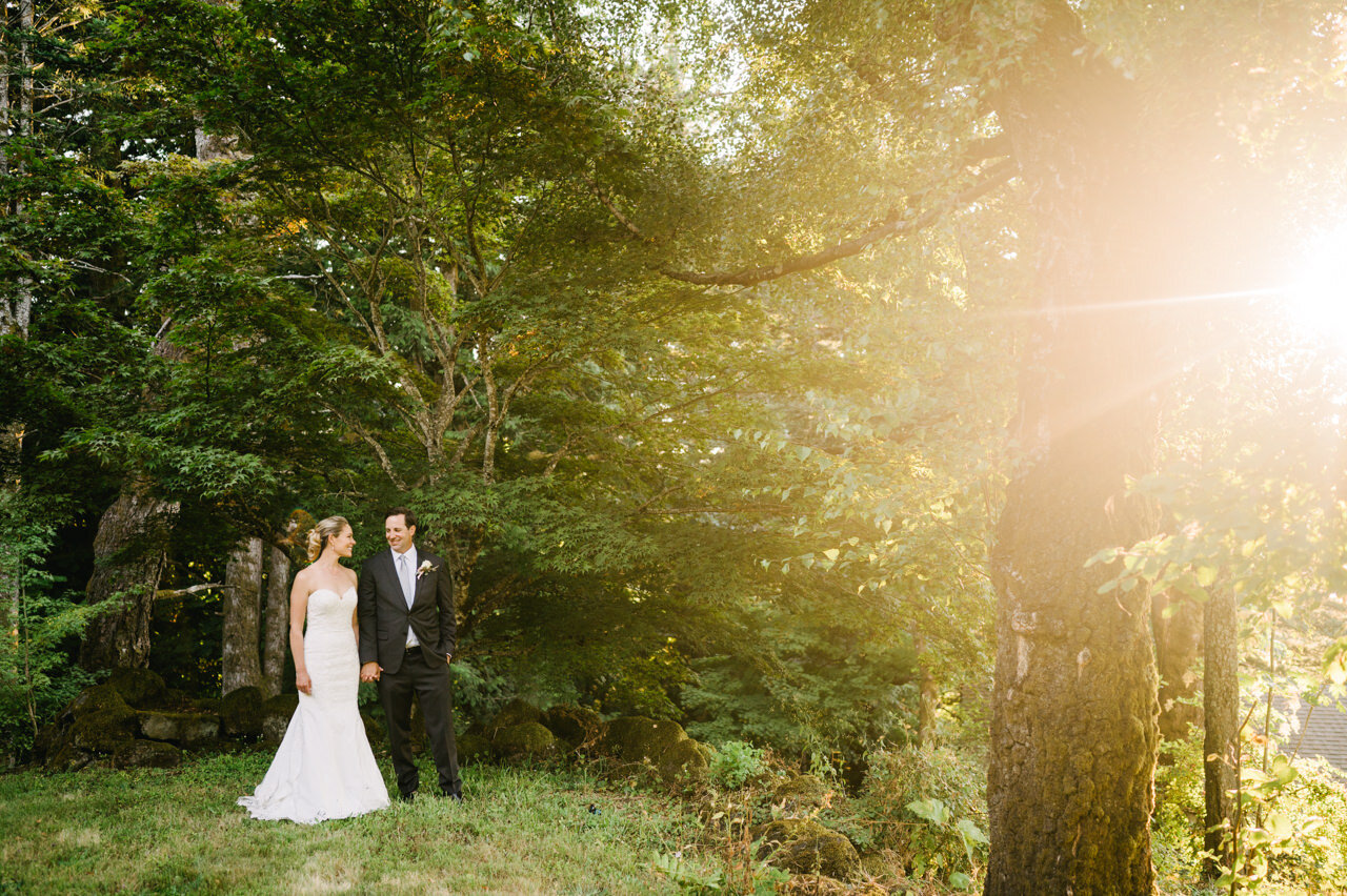  Bride and groom standing to the left while sunlight shines through trees 