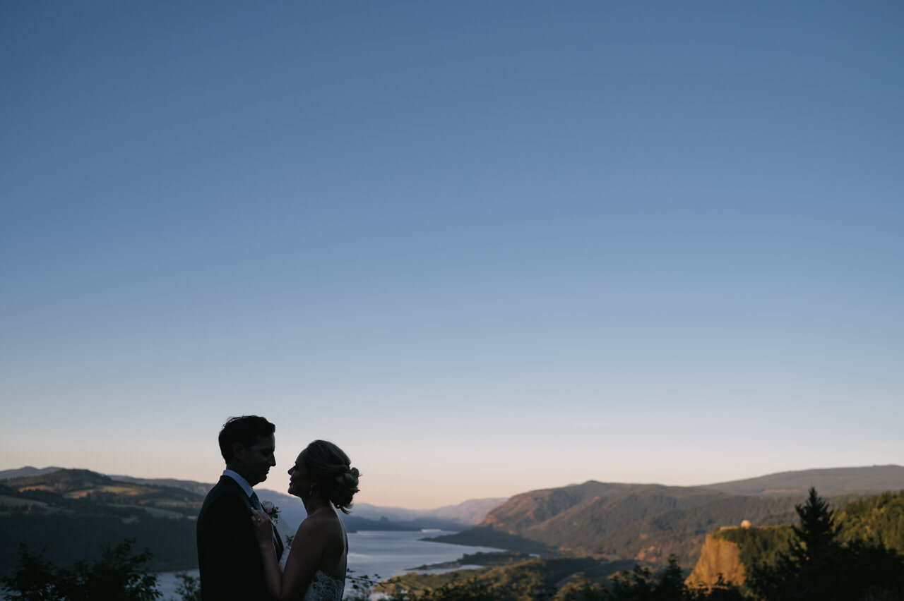  Bride and groom look at each other from menucha retreat center viewpoint of Columbia gorge with vista house behind them 