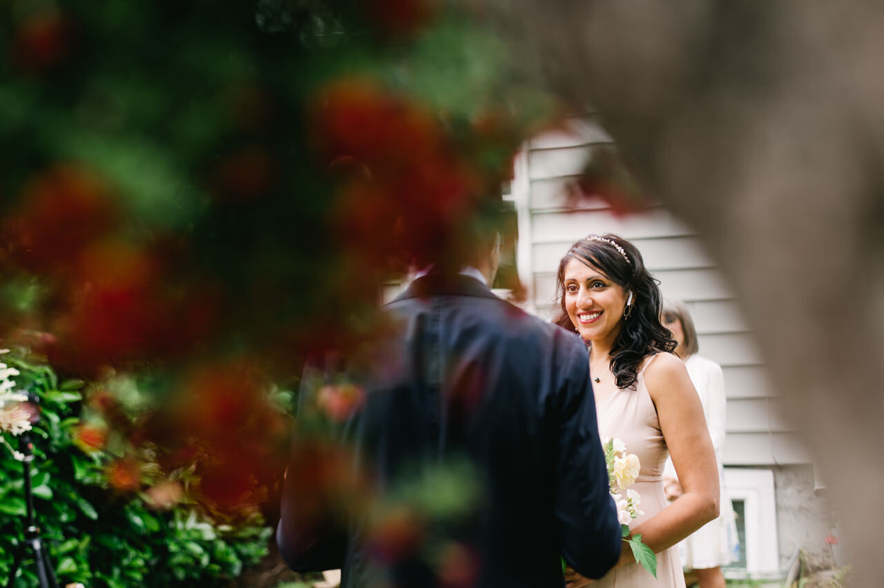  Bride smiles at groom during elopement ceremony with hanging flowers in foreground 