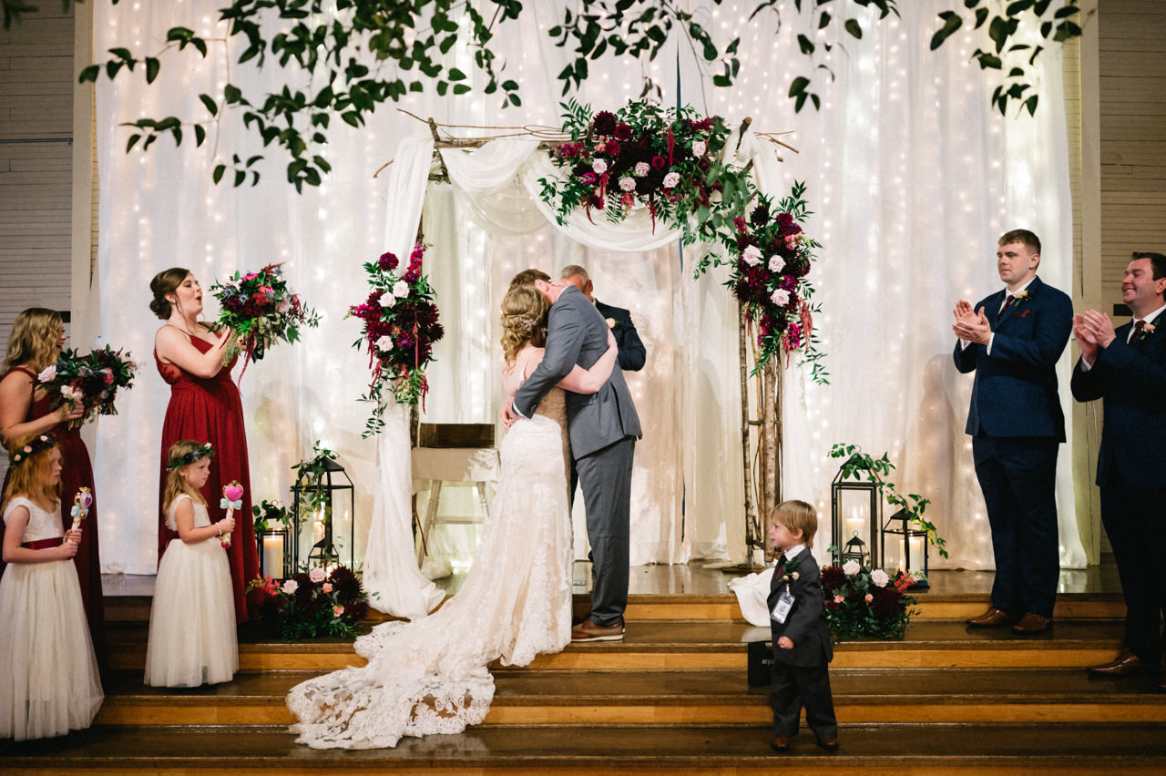  Bride and groom kiss under hanging maroon, pink, and green florals at green villa barn with bridal party cheering 