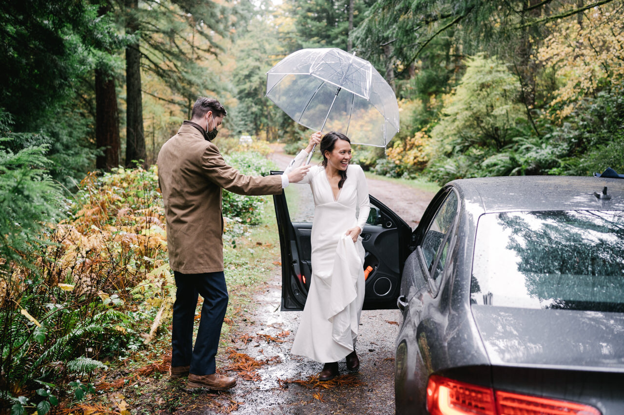  Happy bride getting out of car with clear umbrella in the rain on forest road 