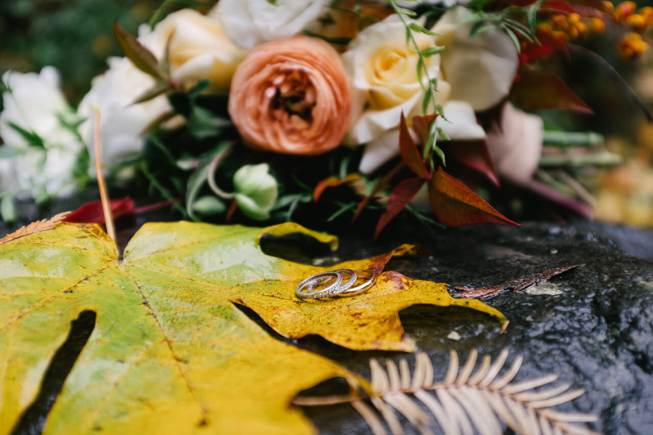  Closeup of wedding rings on fall Maple leaf with orange and white bouquet in background 