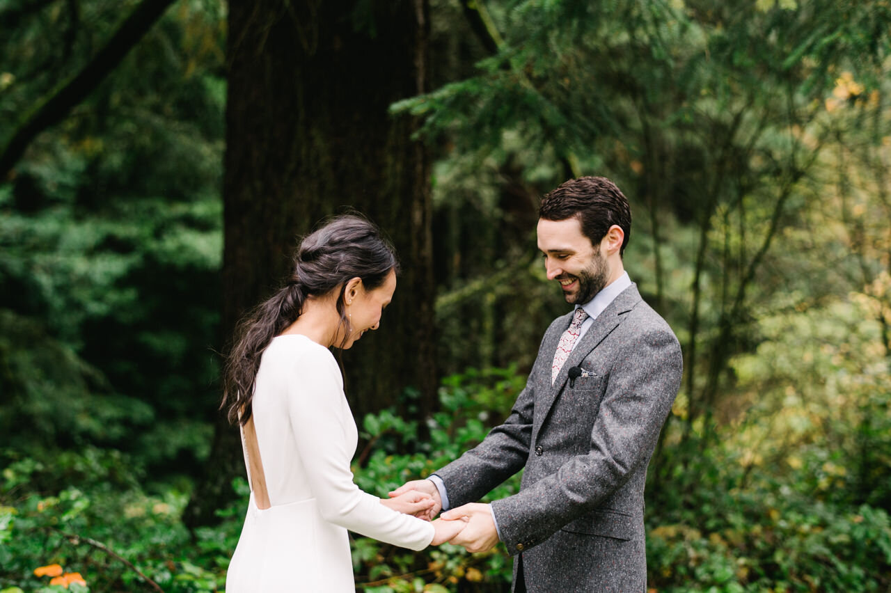  Groom smiles at bride's wedding dress in front of forest 