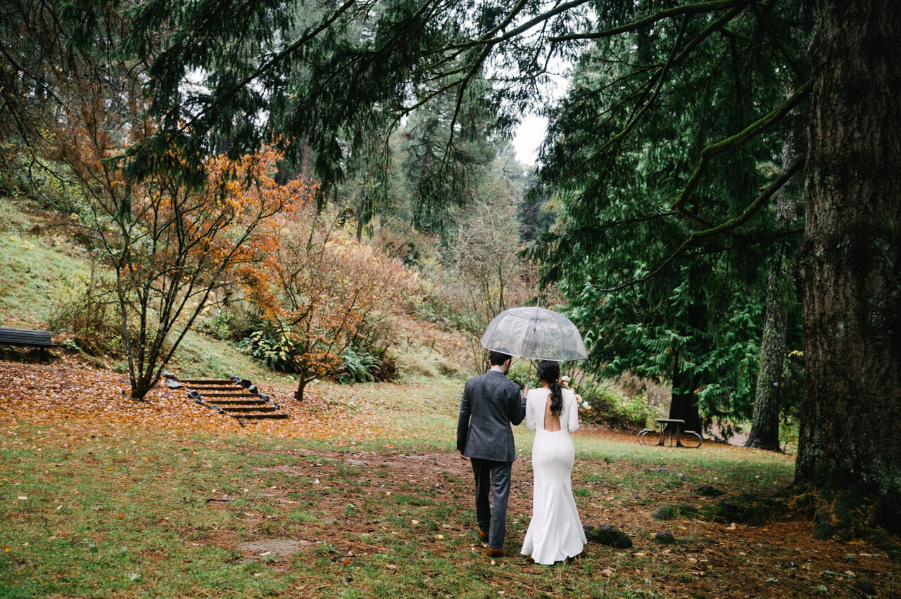  Bride and groom walk away in forest park with umbrellas in rainy fall day 