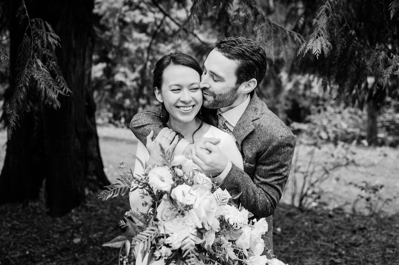  Black and white photo of groom wrapping arms around bride and kissing her cheek 