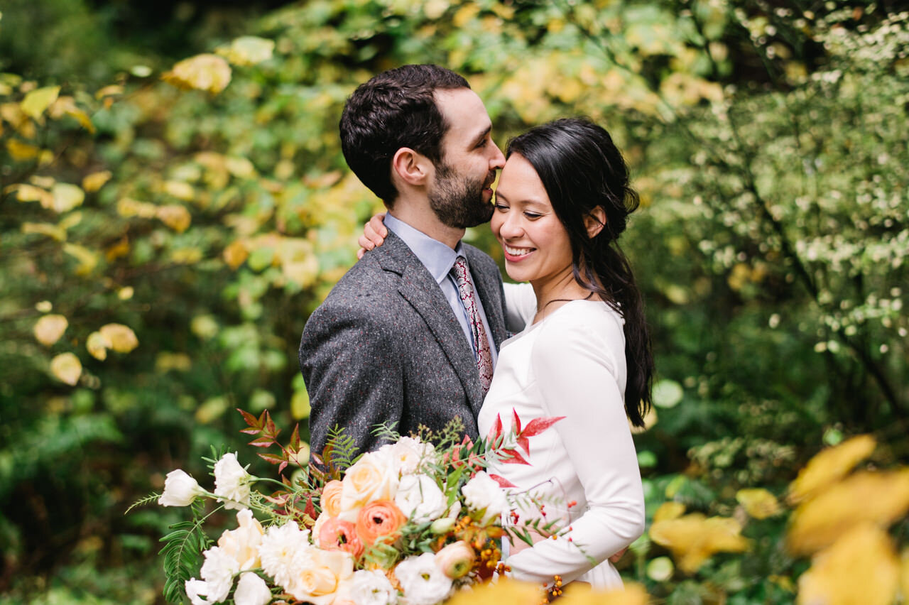  Bride smiling as groom snuggles against her with fall forest yellow leaves wrapped around them 