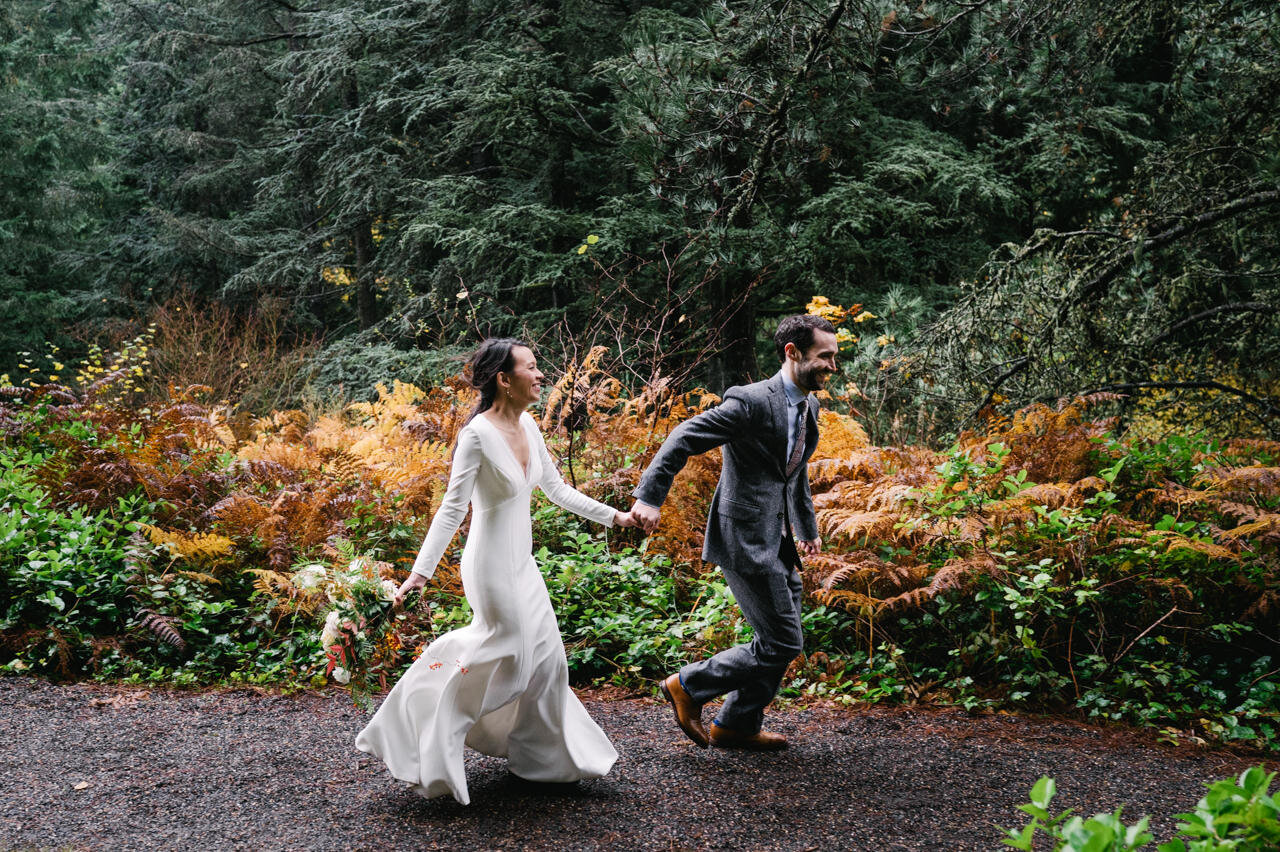  Groom holding hands with bride as they run down forest trail with orange and green ferns behind them 