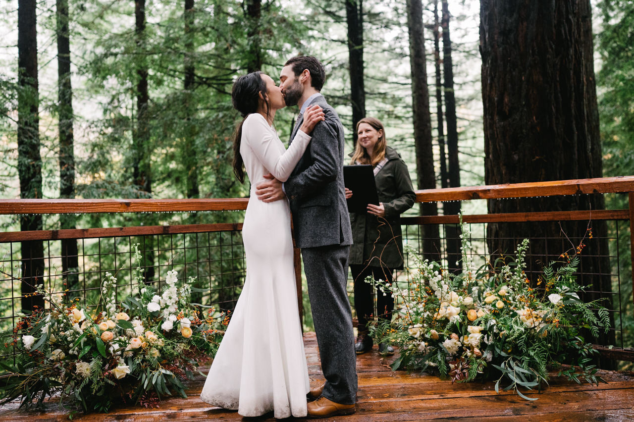  Bride and groom kiss on redwood deck wedding ceremony with large floral centerpieces on each side 